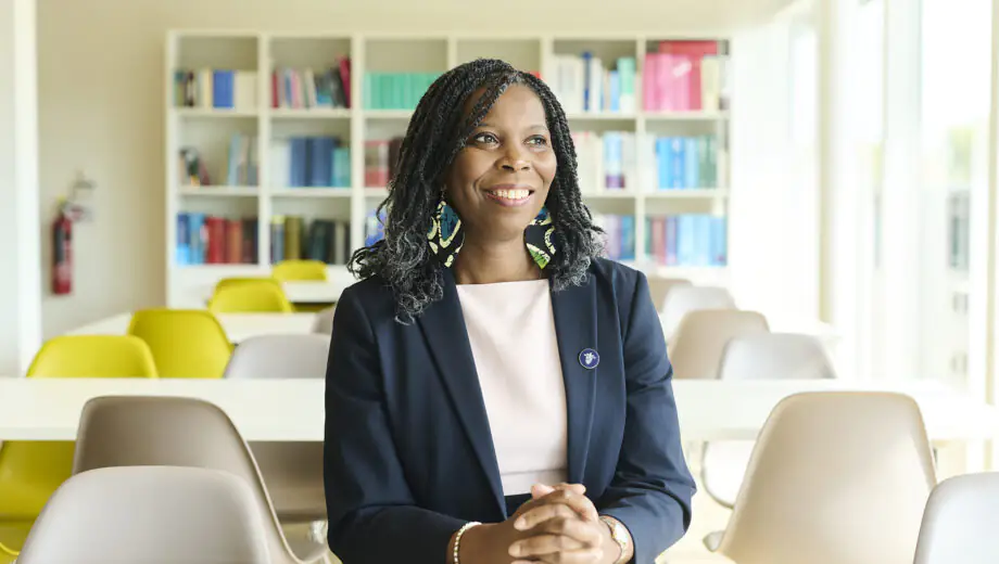 Tase smiles looking into the distance. She wears a white dress and navy blazer with a pin, blue and green earrings. Background is a sunny cafeteria with a bookcase visible in the back