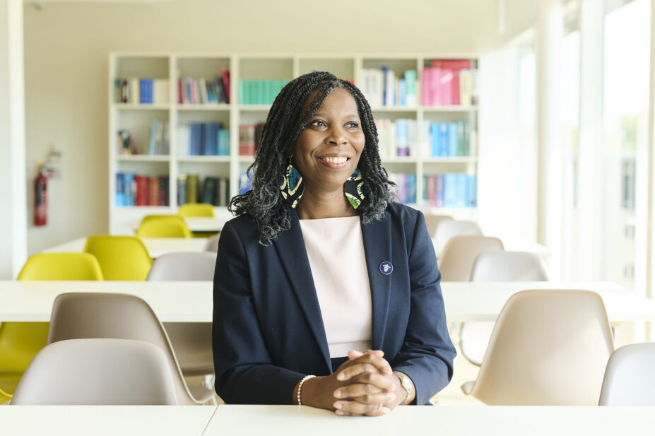 Tase smiles looking into the distance. She wears a white dress and navy blazer with a pin, blue and green earrings. Background is a sunny cafeteria with a bookcase visible in the back