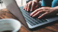 Closeup image of a woman's hands working and typing on laptop keyboard with coffee cup on wooden table