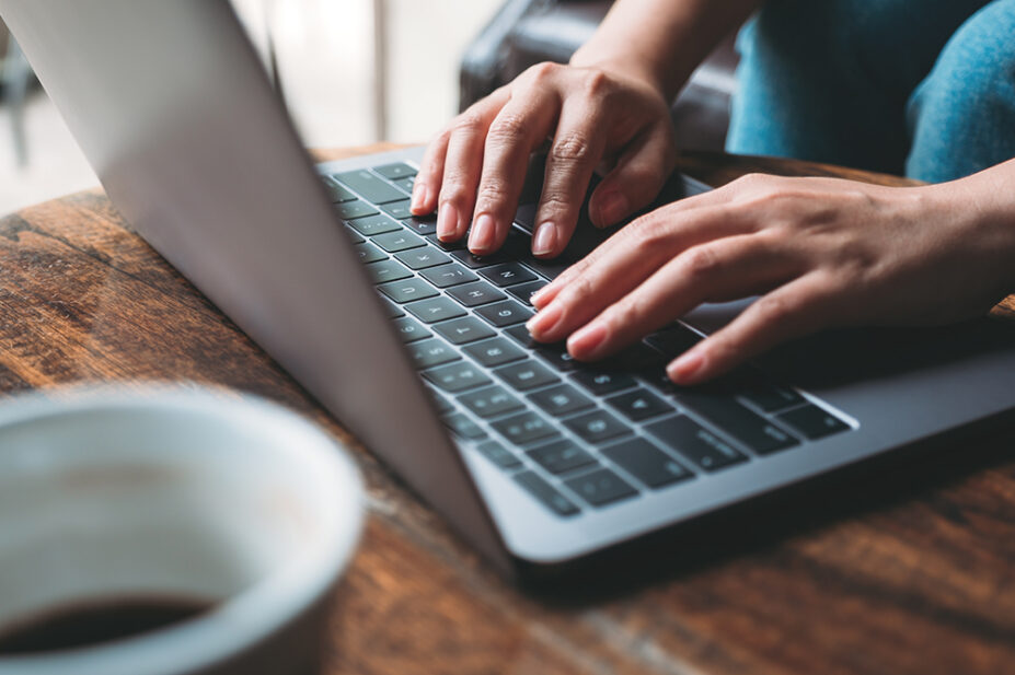 Closeup image of a woman's hands working and typing on laptop keyboard with coffee cup on wooden table